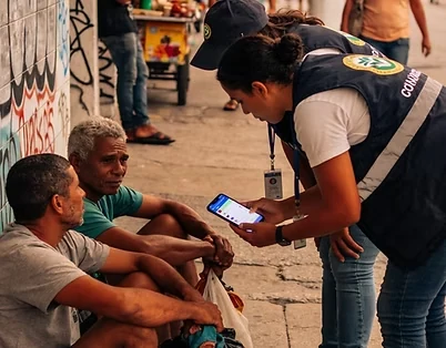 Agente de abordagem social, de colete, consulta o celular ao lado de dois homens sentados na calçada