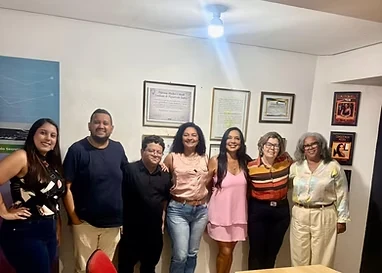 Grupo de sete pessoas posando em uma sala com quadros e certificados na parede